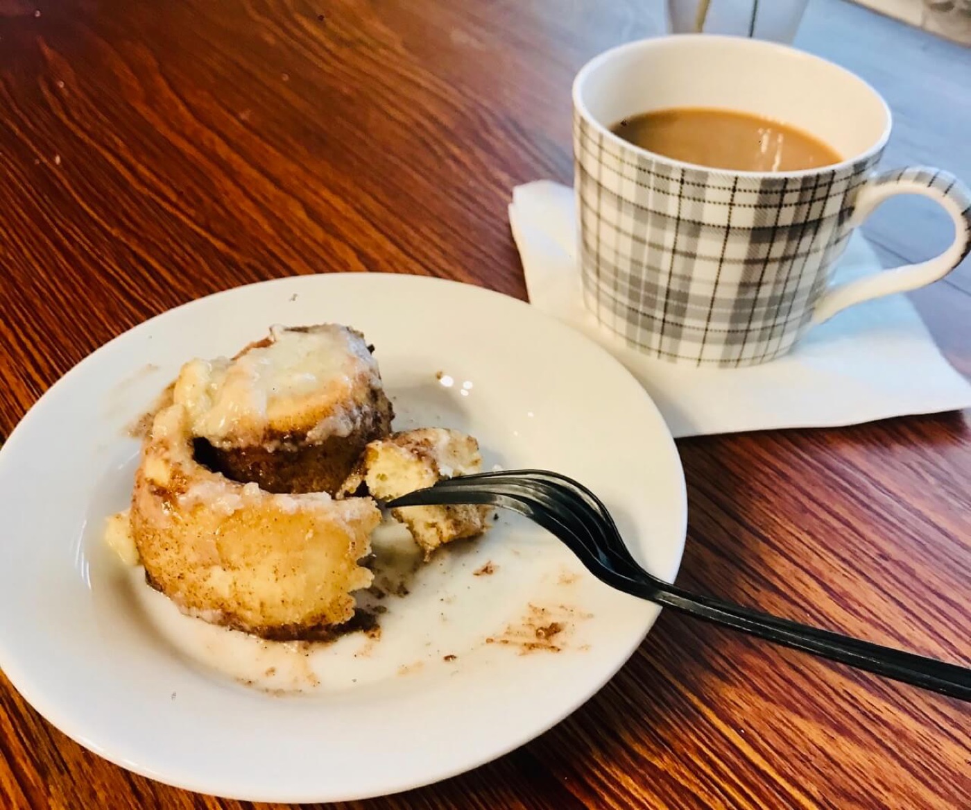 A cinnamon roll on a white plate with a fork and a mug of coffee