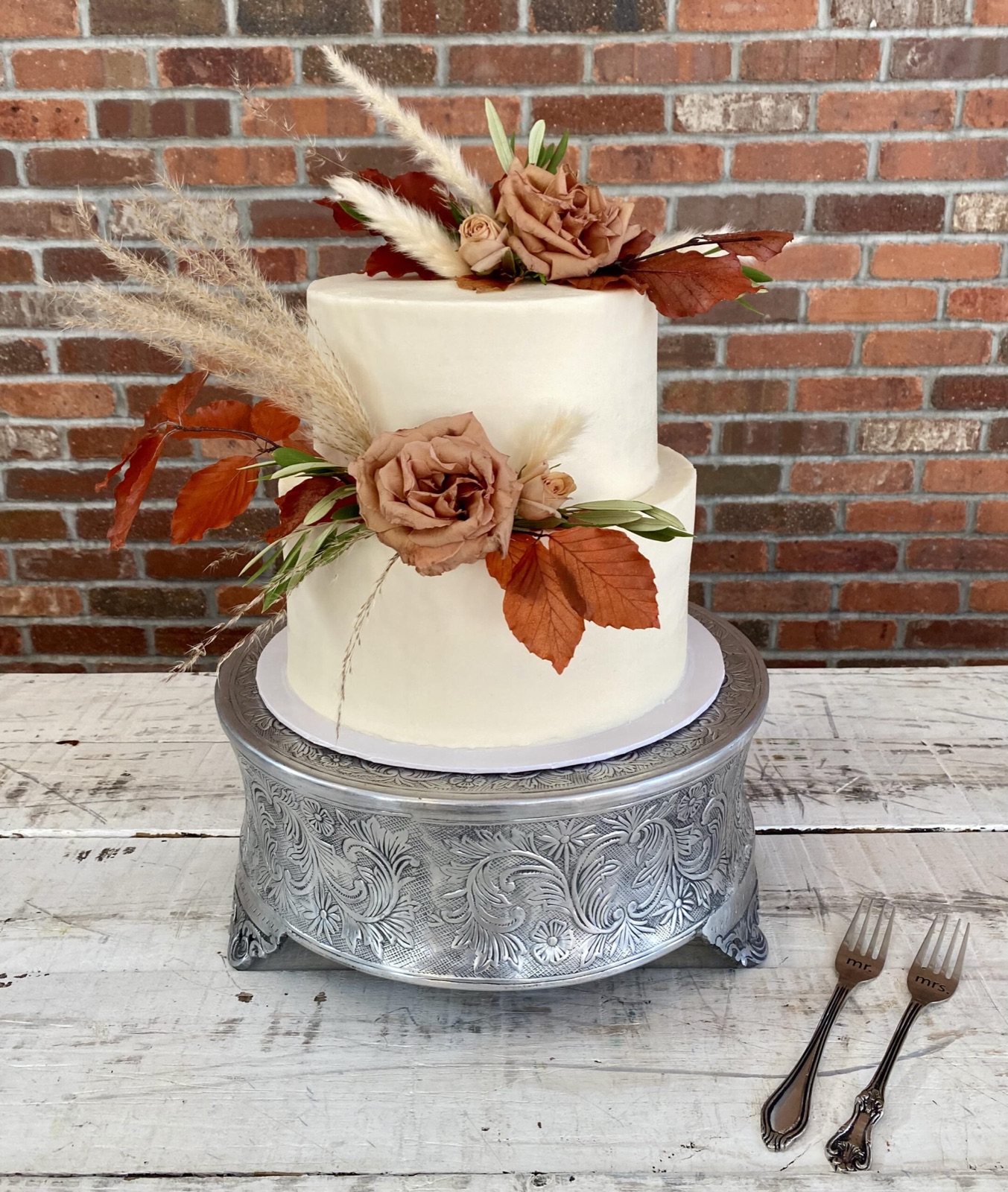A two-tier white buttercream cake with dried florals on a galvanized stand against a brick wall